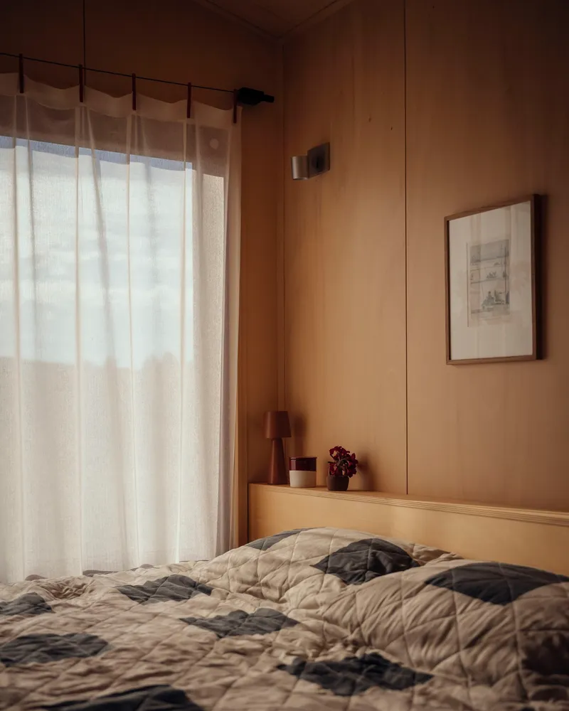 Bedroom corner with unmade bed beneath sheer curtains filtering soft daylight through the plywood-paneled walls