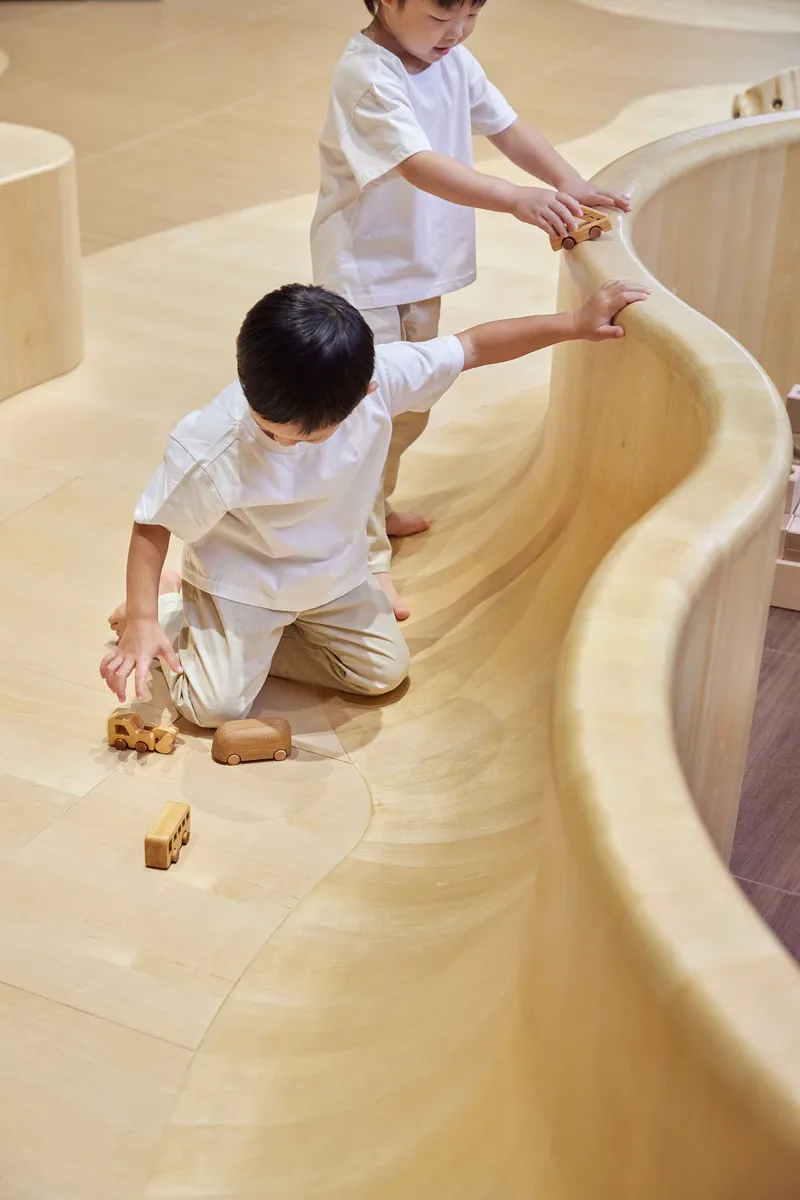 Two children playing with wooden toys along the edge of a curved plywood barrier