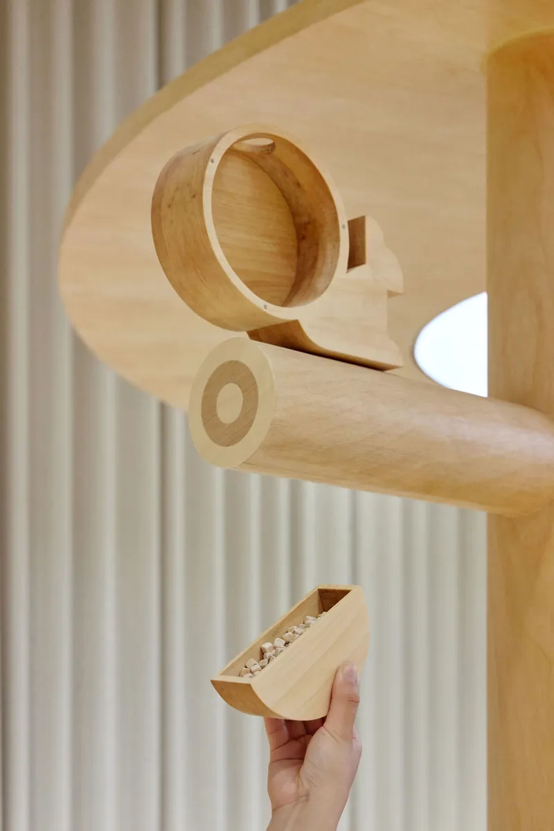 Hand holding a wooden toy piece beneath a cylindrical timber drum suspended from plywood structure