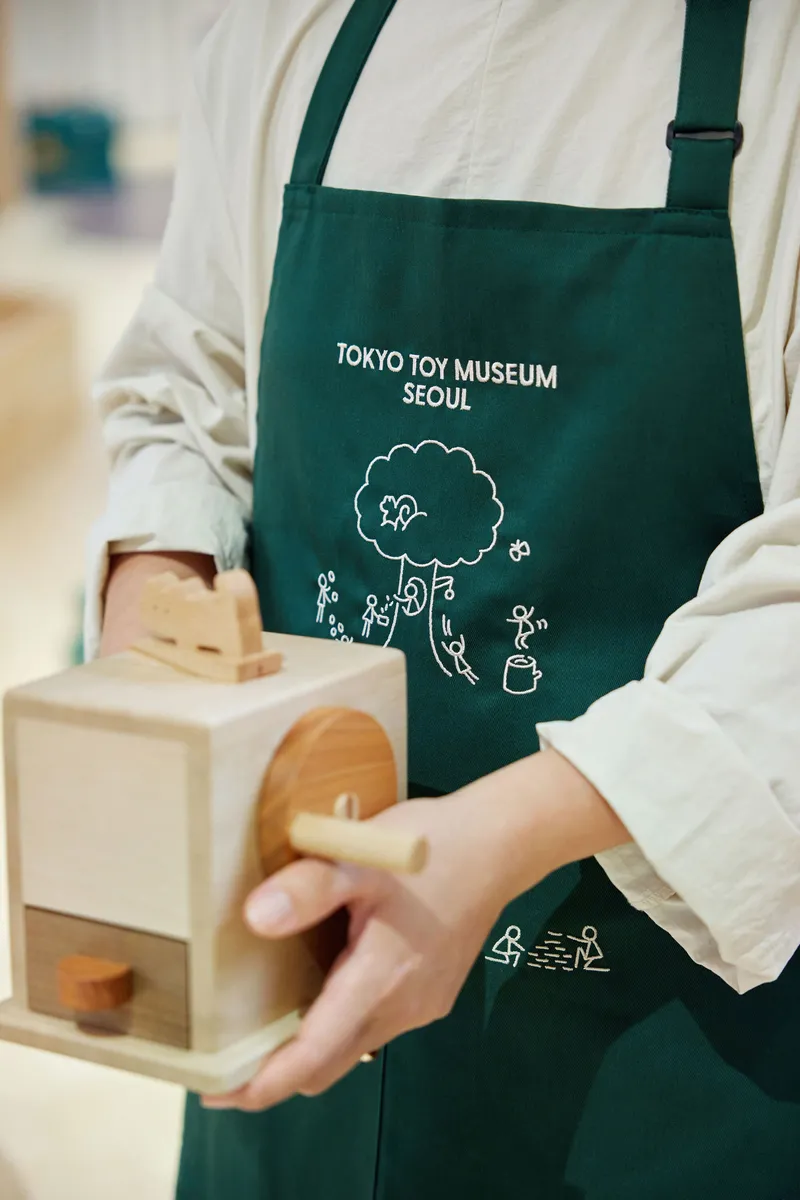 Close-up of hands holding a wooden toy model while wearing a green apron with logo