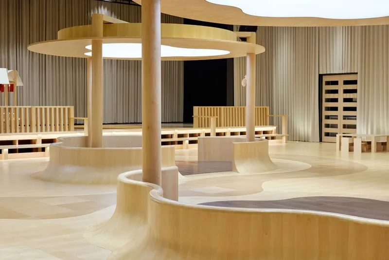 Curved plywood benches and platforms surrounding cylindrical columns beneath ring-shaped pendant lights