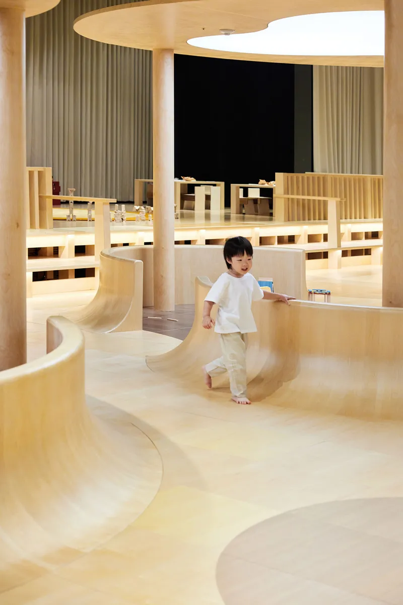 Curved plywood ramps and platforms beneath a suspended ceiling canopy with a child descending