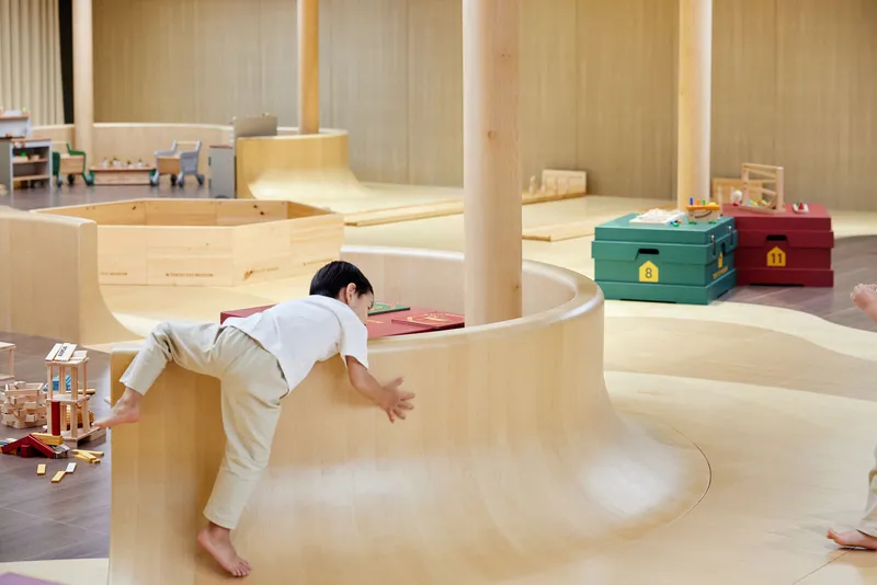 Child climbing over a curved plywood partition with toys scattered on the floor below