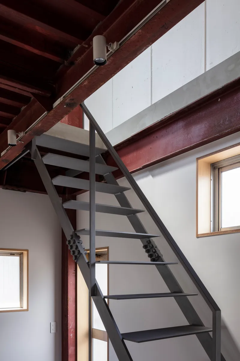 Steel stair rising past plywood-framed windows beneath the exposed timber ceiling structure