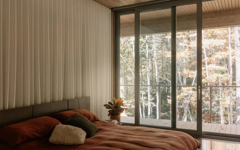 Bedroom with floor-to-ceiling glazing opening to a balcony overlooking autumn trees in soft morning light