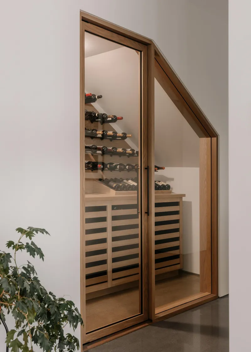 Angled glass door framing a wine cellar with horizontal slat cabinet and bottle racks beside a potted plant