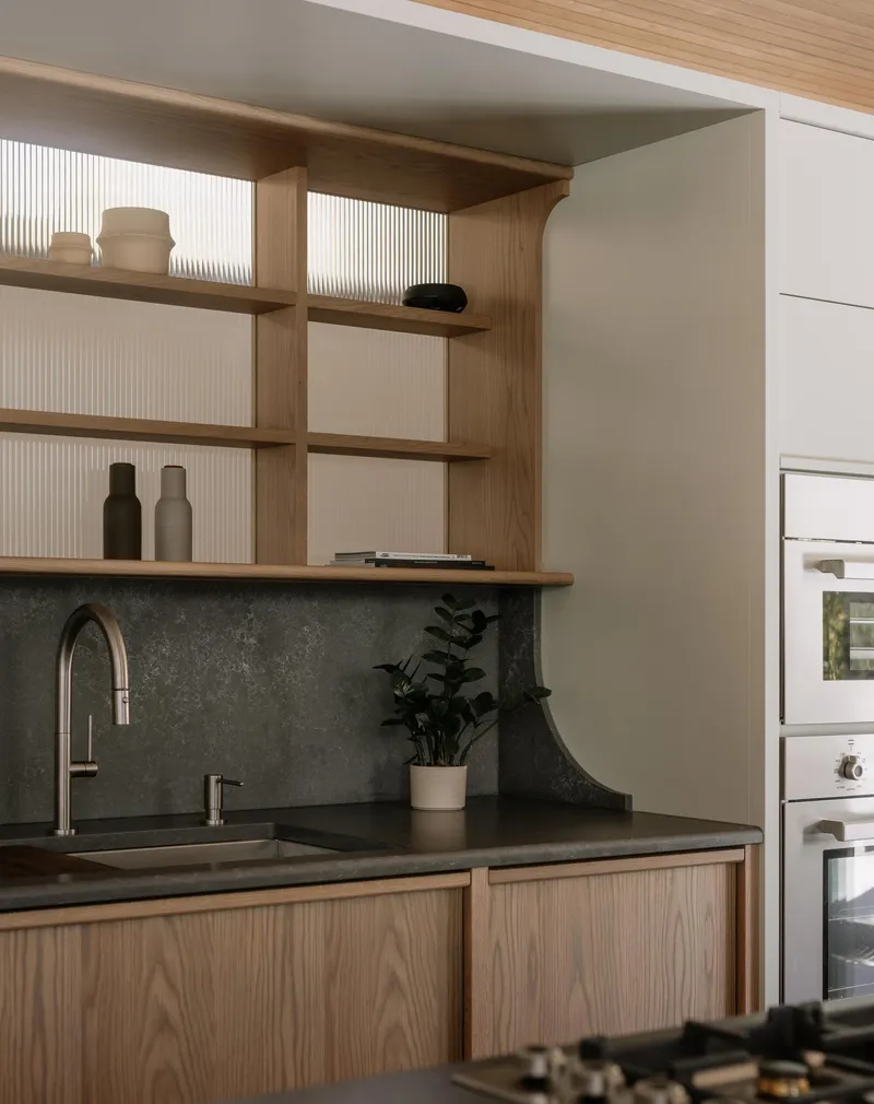 Kitchen counter with timber shelving, fluted glass panels, and dark stone backsplash under natural light