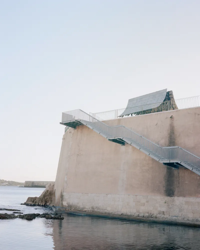 Metal staircase ascending the exterior concrete wall above rocky shoreline water