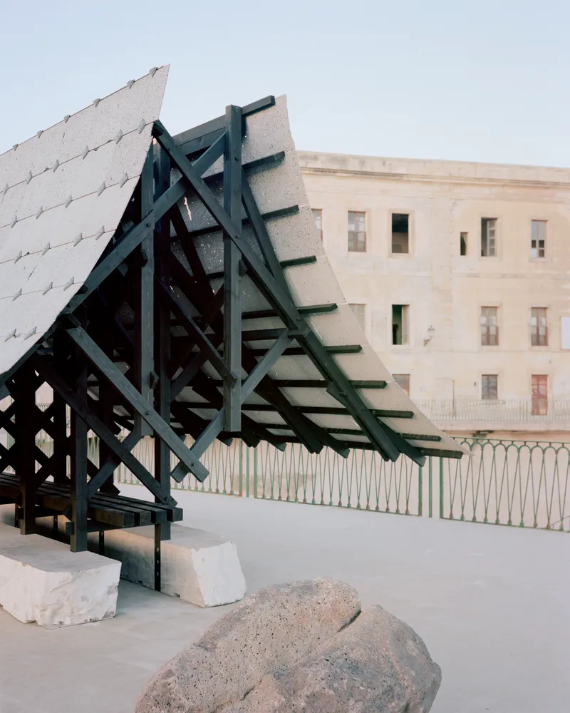 Angled timber pavilion with exposed structural frame and stone cladding set against a three-story stucco building