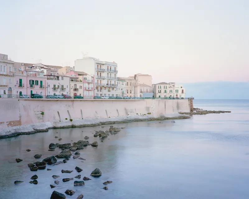 Waterfront residential buildings at dusk with exposed rocks in the shallows below the seawall
