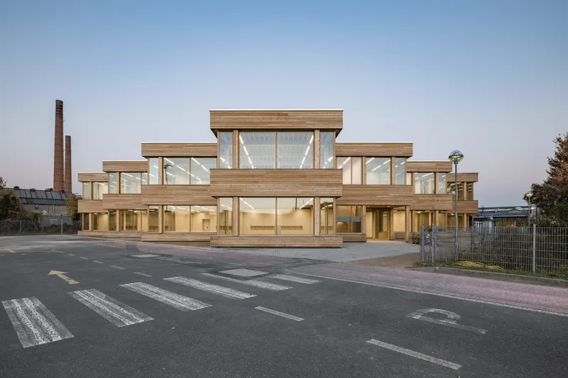 Stepped timber and glass facade fronting a crosswalk at dusk with brick chimneys beyond