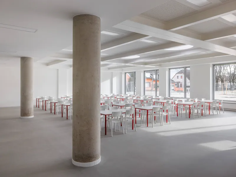 Interior dining hall with cylindrical concrete columns and rows of red and white chairs below a coffered ceiling