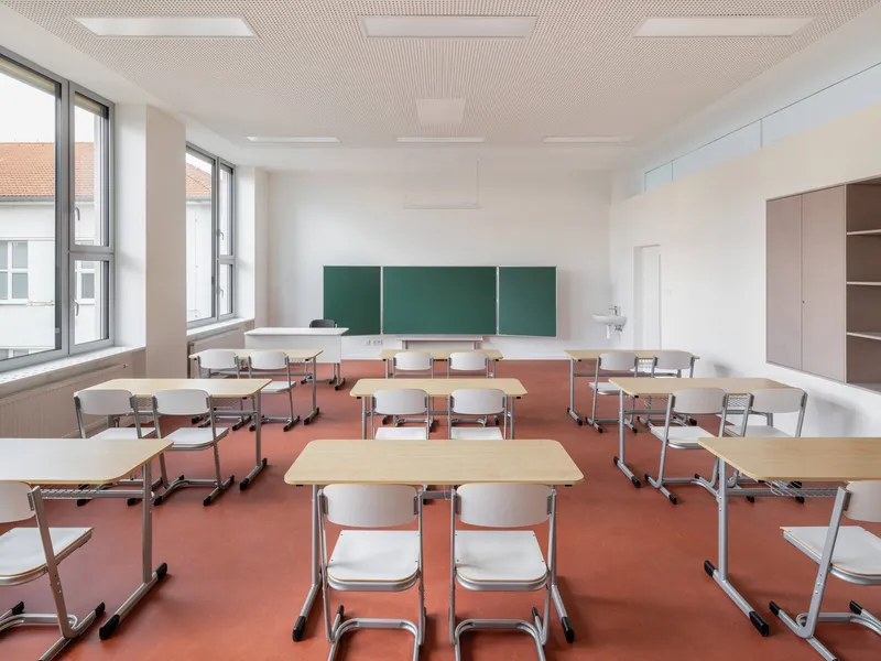 Classroom interior showing rows of movable desks and stacking chairs arranged on the terracotta floor facing the chalkboard