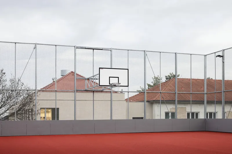 Rooftop sports court with red surface and basketball hoop behind wire mesh fencing under overcast sky