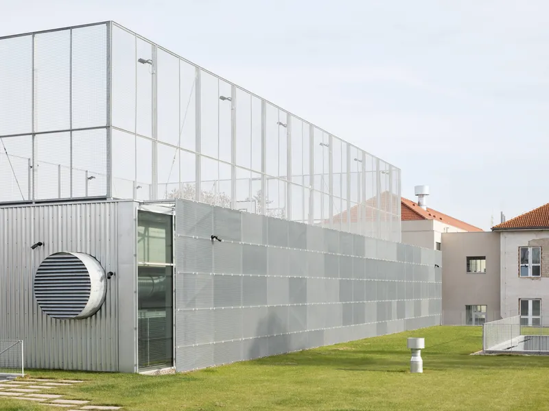 Exterior facade with corrugated metal panels featuring circular window and translucent mesh screen under grey clouds