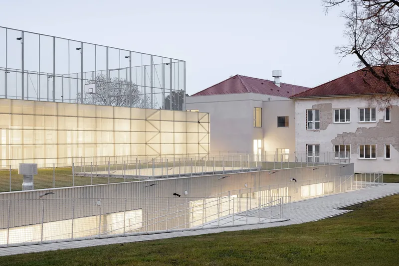 Translucent polycarbonate wall glowing at dusk beside an existing residential building with tiled roof