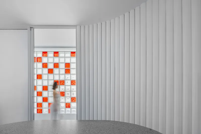 Interior view of a white vertical panel wall framing a grid window with orange and white glass blocks