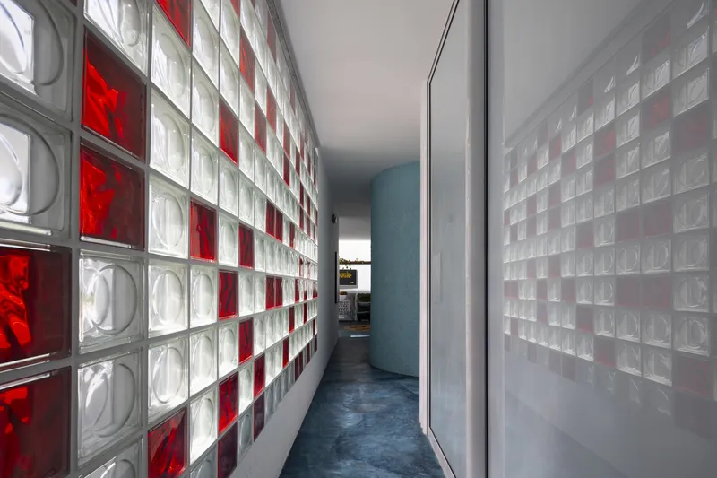 Narrow passage flanked by red and clear glass block walls with blue concrete floor extending toward daylight