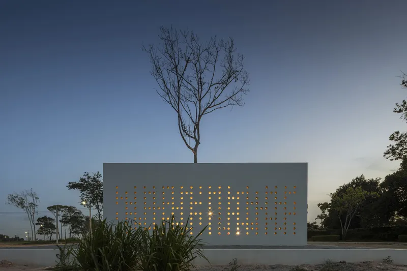 Perforated concrete facade with illuminated openings and tree emerging from rooftop at dusk