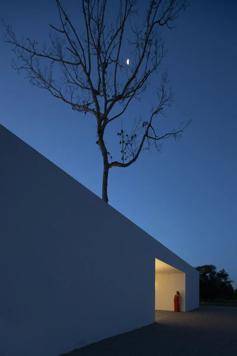 Angled white wall with glowing doorway where a figure in red stands under a bare tree at twilight