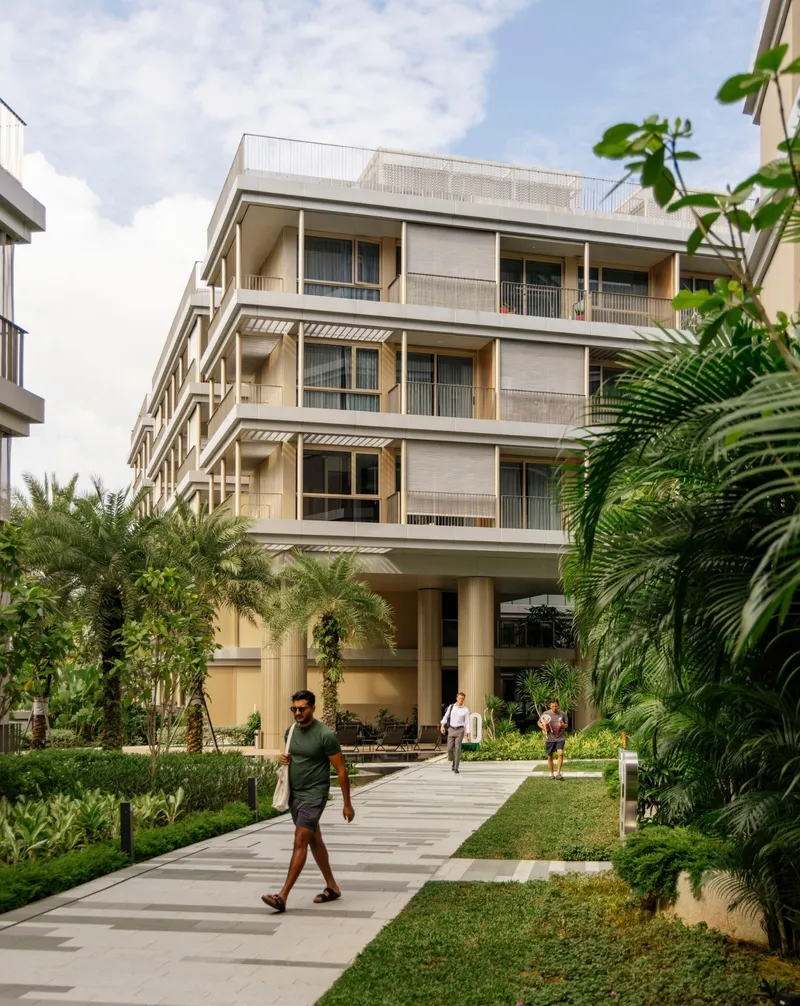 Courtyard pathway through palms and planted beds toward an elevated residential block with people walking in daylight