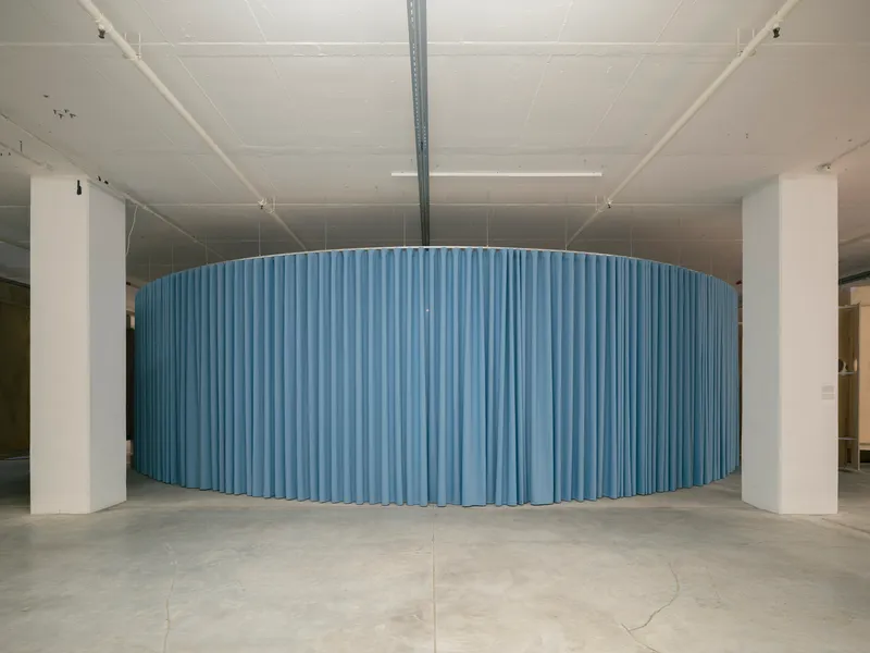 Curved blue corrugated metal wall spanning between white columns in gallery space with polished concrete floor