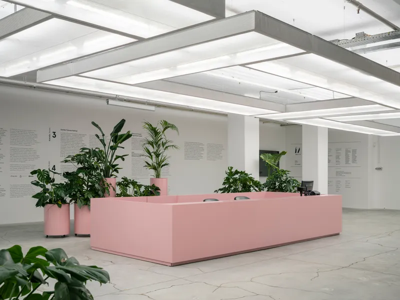 Pink reception desk with cylindrical planters and tropical foliage beneath white coffered ceiling with linear lighting