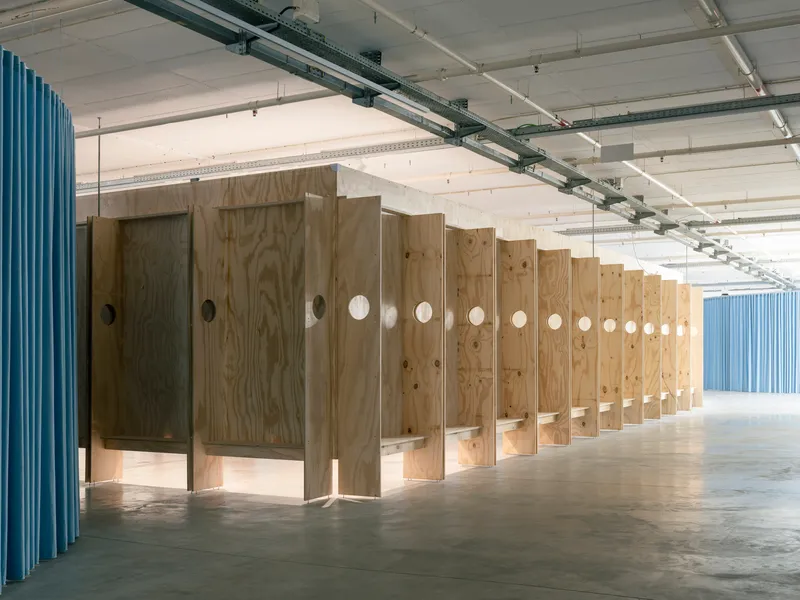 Row of plywood partitions with circular openings flanked by blue corrugated walls under exposed concrete ceiling