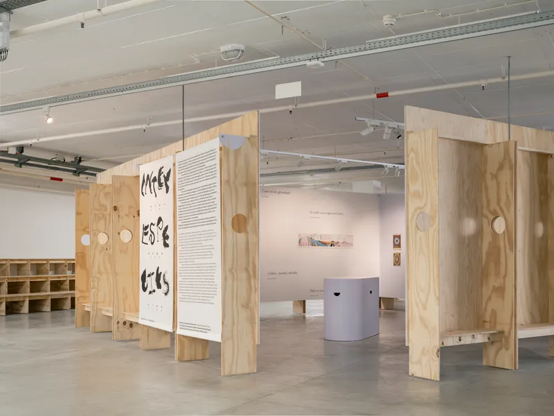 Gallery interior with freestanding plywood partition walls featuring circular cutouts and text panels under exposed ceiling