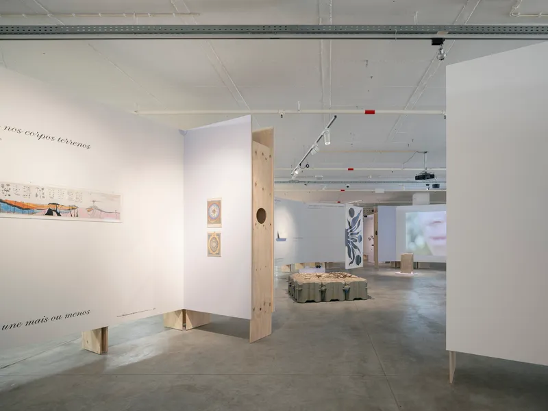 Exhibition space showing white display walls and plywood elements with artworks and projection visible in background