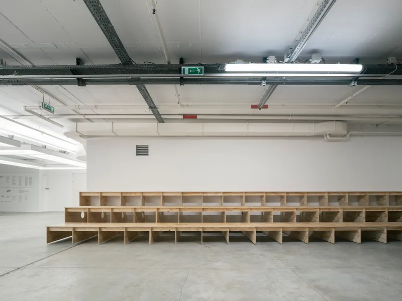 Long tiered plywood seating structure with stepped risers spanning the width of a white gallery space