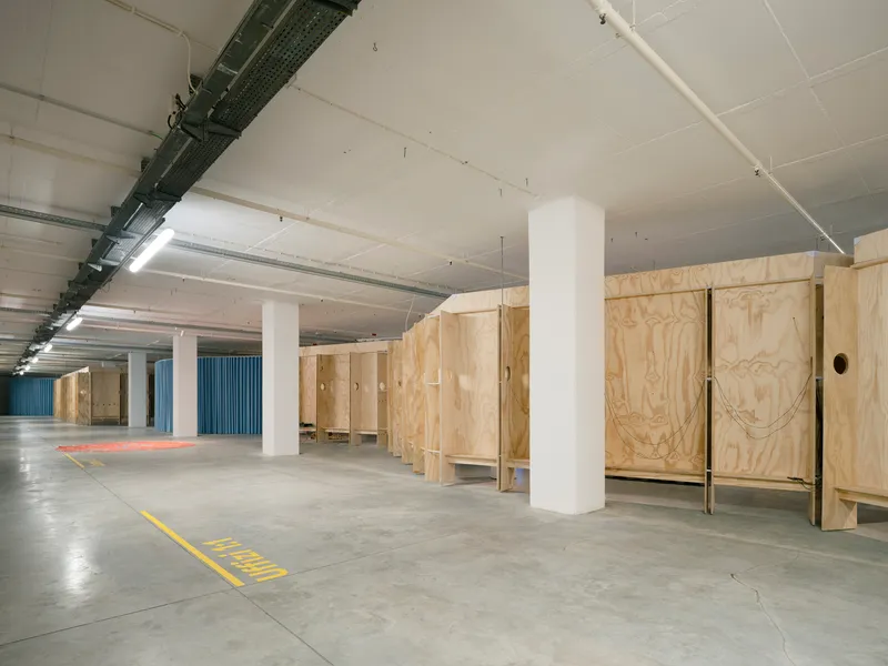 Open gallery space with white cylindrical columns and freestanding plywood storage units under fluorescent lighting