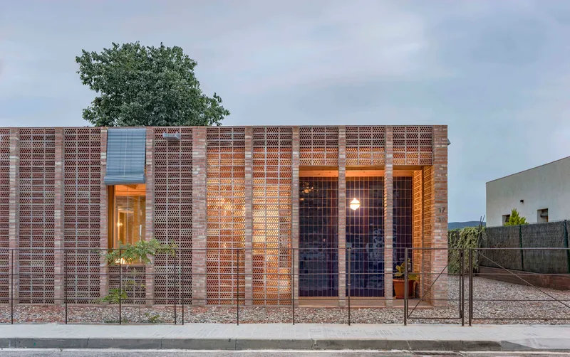 Street facade at dusk showing illuminated interior through perforated brick and steel mesh screens with tree above