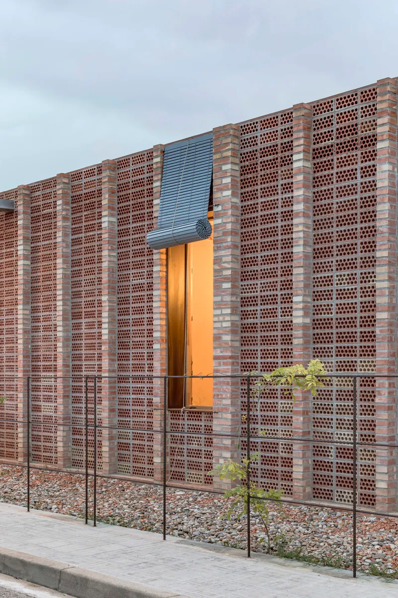 Corner detail of perforated brick wall with steel mesh panel and illuminated doorway at twilight