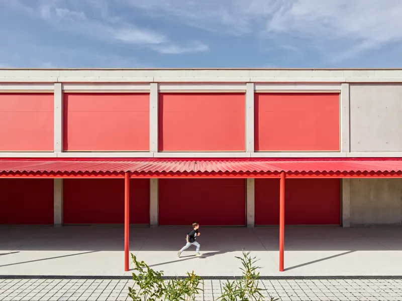 Red corrugated canopy supported by steel columns sheltering a paved walkway with a running child