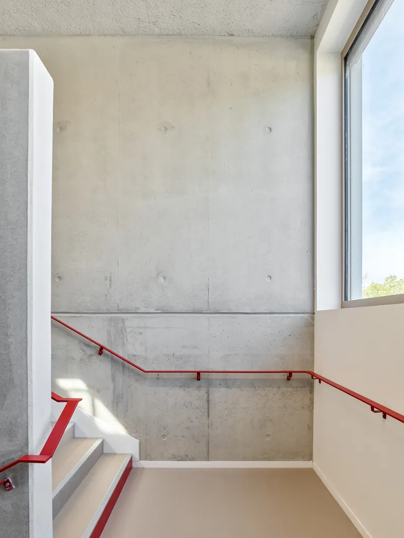 Stairwell with cast concrete walls, red steel handrail and natural light from a corner window