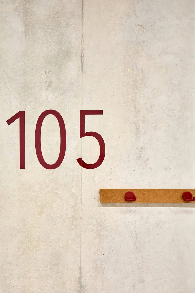 Room number 105 in maroon typography on a board-formed concrete wall with timber coat hooks