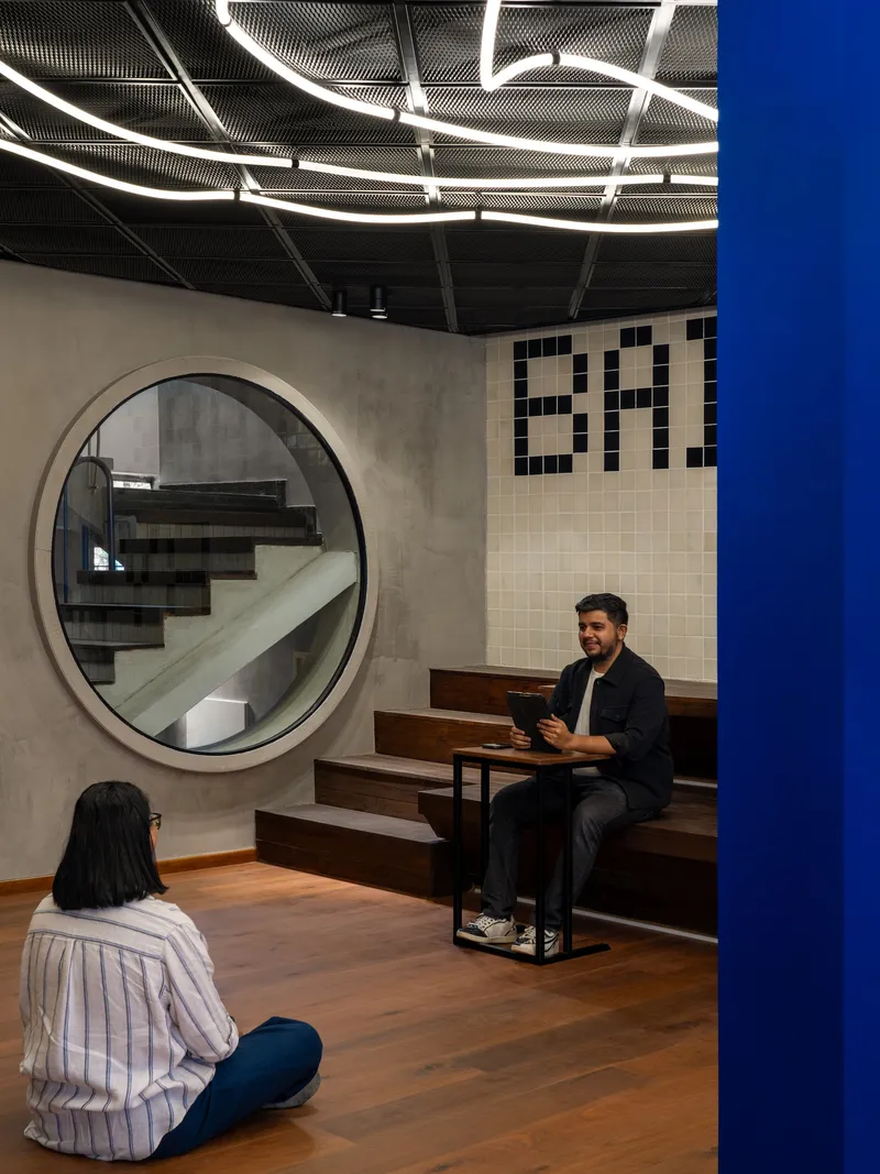 Seating area with circular concrete staircase portal, tiled wall graphics, and two people beneath curved linear lighting