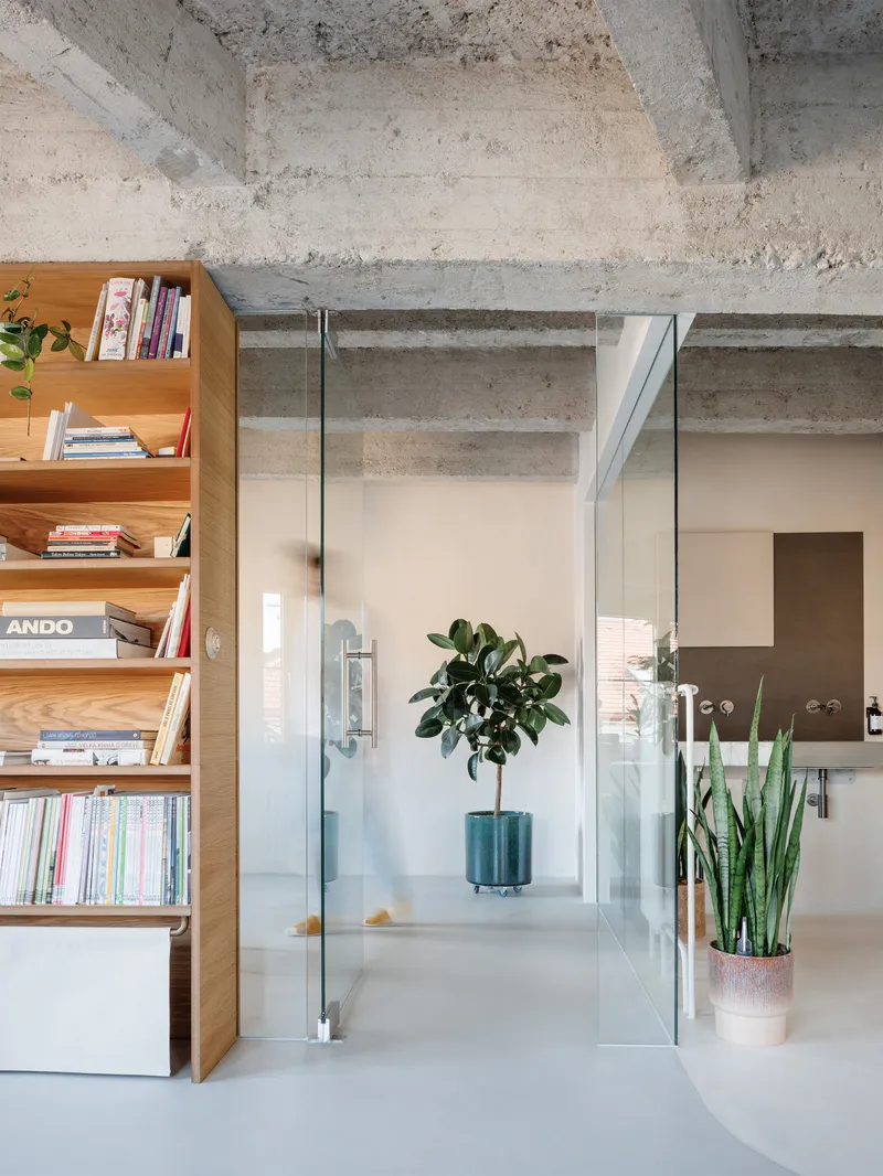 Glass-enclosed room viewed through open door with timber bookshelf, potted plants, and exposed concrete structure