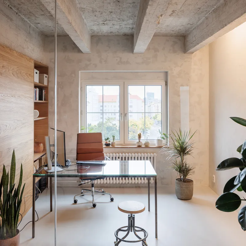 Office space with exposed concrete ceiling beams, glass desk and potted plants by the window