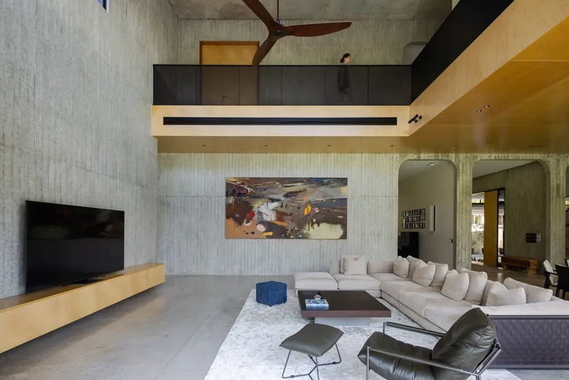 Double-height living room with board-formed concrete walls, exposed timber beams, and timber-lined mezzanine above