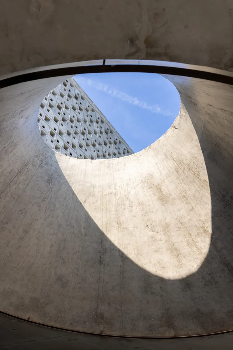 View upward through a teardrop-shaped concrete oculus framing perforated facade and blue sky