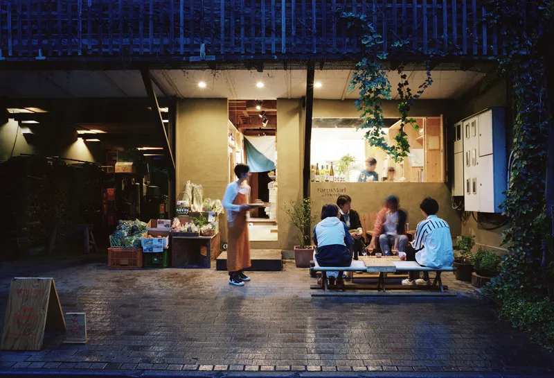 Glass-fronted shopfront opening to a paved courtyard with people gathered at dusk
