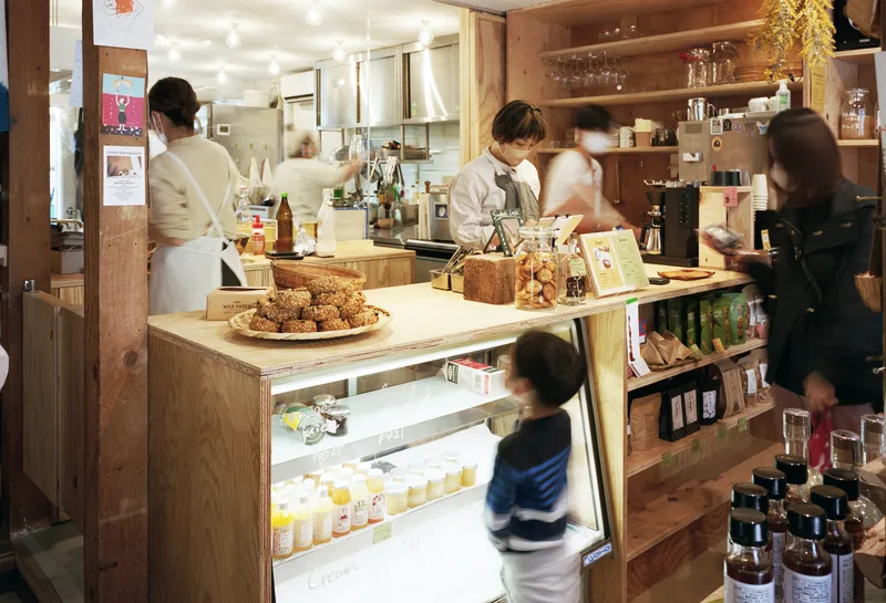 Plywood service counter with display case and open shelving as staff prepare food in the kitchen beyond