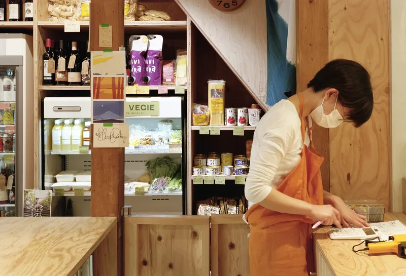 Staff member at plywood counter with open shelving displaying packaged goods and refrigerated case