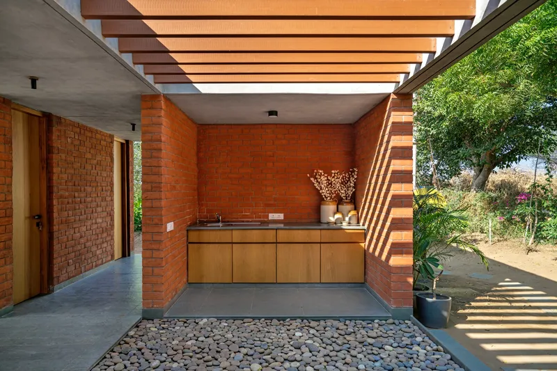 Semi-outdoor kitchen counter beneath timber pergola slats with river stone flooring and afternoon shadows