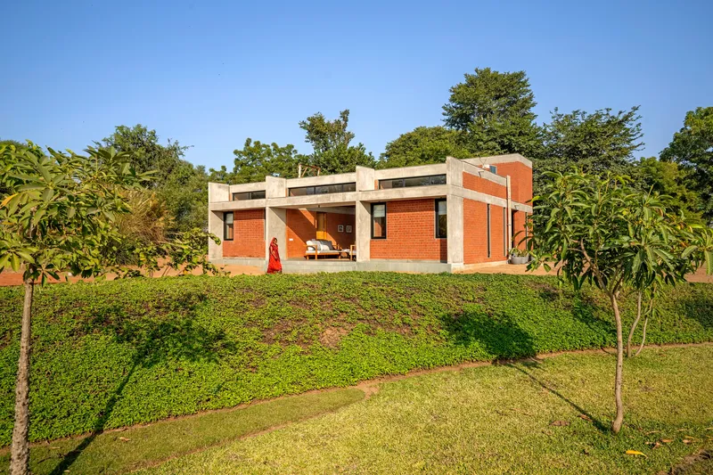 Red brick residence atop a grassed slope with scattered young trees under clear blue sky