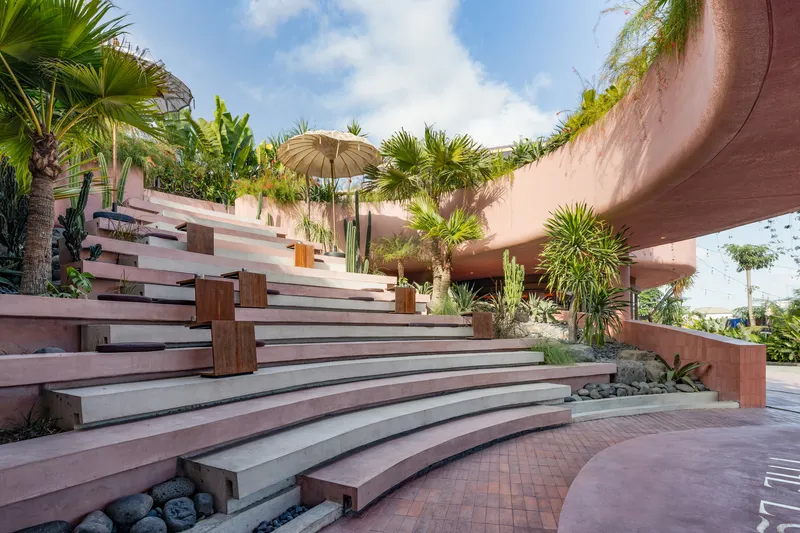 Wide terraced amphitheater steps in pink and grey tones surrounded by palms and cacti under a partly cloudy sky