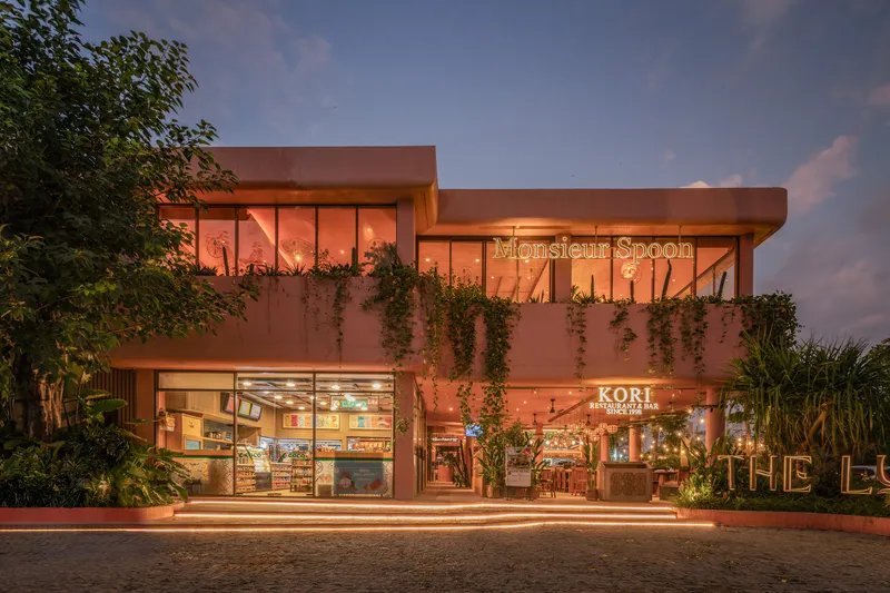 Street view of the two-story terracotta facade with planted balconies and retail storefronts at dusk