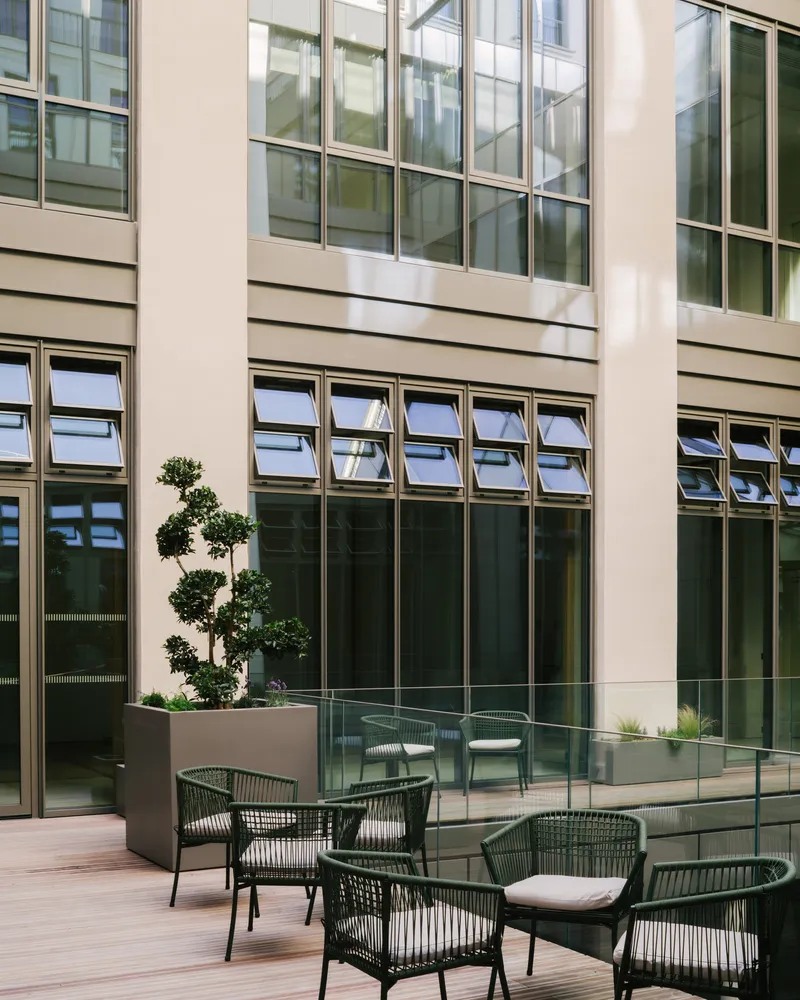 Courtyard terrace with louvered windows, glass balustrade, topiary tree in planter, and woven outdoor chairs